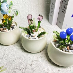 Puede incluir: Tres pequeñas macetas de cerámica blanca con arreglos florales de vidrio. Las macetas están llenas de pequeñas piedras blancas. Las flores son amarillas, rosas y azules.