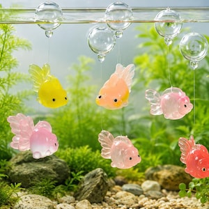Puede incluir: Seis figuras de peces dorados translúcidos en colores amarillo, melocotón, rosa y rojo, cada una suspendida por un hilo fino y transparente de una pequeña esfera de cristal que flota en la superficie del agua. Tienen aletas y ojos negros.