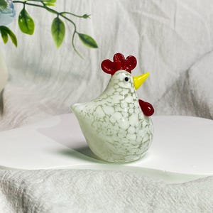 May include: White glass chicken figurine with red and yellow accents. The chicken is standing on a white surface.
