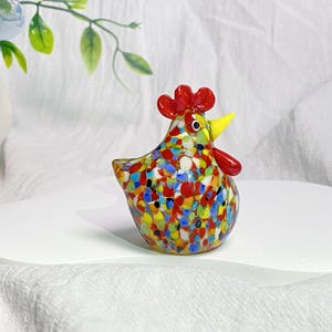 May include: A colorful glass chicken figurine with red, yellow, blue, and white speckled paint. The chicken has a yellow beak and red comb.