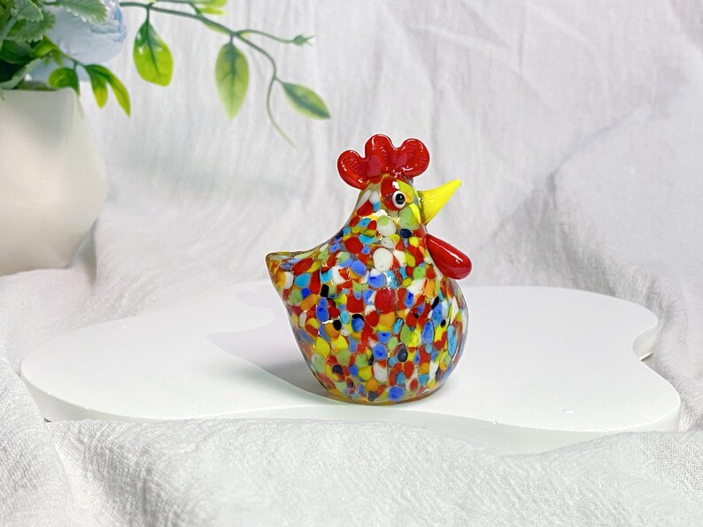May include: A colorful glass chicken figurine with red, yellow, blue, and white speckled paint. The chicken has a yellow beak and red comb.