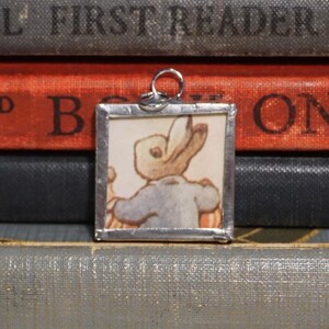 May include: A square pendant with a silver-colored frame features a vintage illustration of a rabbit wearing a blue jacket. The pendant hangs from a silver-colored loop. The background consists of stacked books with red and black covers.