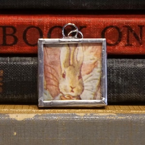 May include: A square pendant with a silver-colored frame features a vintage illustration of a rabbit. The rabbit has long ears and is wearing a light-colored jacket. The pendant hangs from a silver-colored loop. The background includes a stack of books.