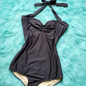 May include: A black halter-neck swimsuit with a sweetheart neckline and a tie at the back. The one-piece swimsuit features a gathered bust and a high-cut leg. The interior lining is a light beige colour. The swimsuit is displayed on a turquoise faux fur surface.
