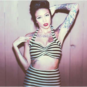 May include: A woman with a black and white striped swimsuit and tattoos on her arms poses in front of a pink wood panel wall. She has red lipstick and her hair is styled in a bouffant.
