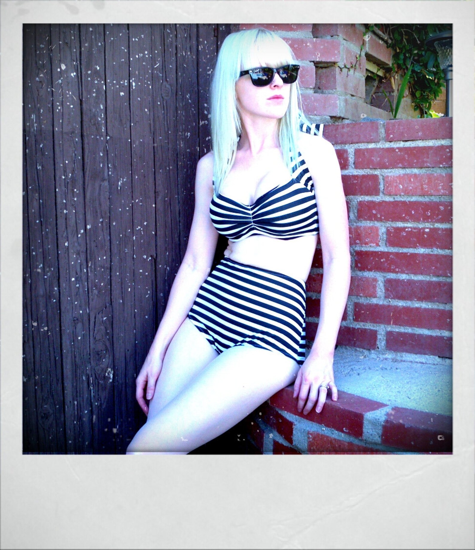 Pale woman in sunglasses wearing a retro goth bikini with black and white stripes, halter top, and high-waisted bottoms, sitting by a brick wall.