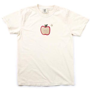 May include: A white t-shirt featuring a red apple graphic and a small gold star. The apple has the word "good" written inside.