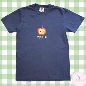 Może przedstawiać: Granatowa koszulka z nadrukiem czerwonego i żółtego jabłka oraz napisem "apple" w kolorze białym. Koszulka ma okrągły dekolt i krótkie rękawy. Materiał wydaje się miękki.