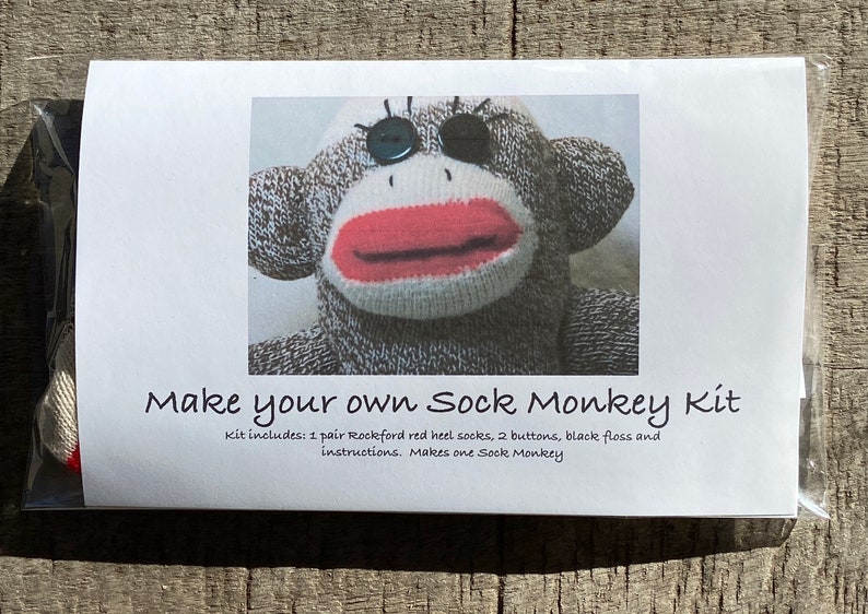 Puede incluir: Una tarjeta blanca con el texto "Make your own Sock Monkey Kit" y una foto de la cara de un mono de calcet&iacute;n. El texto debajo de la foto dice "Kit includes: 1 pair Rockford red heel socks, 2 buttons, black floss and instructions. Makes one Sock Monkey."