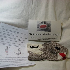 Puede incluir: Un kit de artesan&iacute;a de mono de calcet&iacute;n con un calcet&iacute;n marr&oacute;n y blanco, botones negros, hilo rojo e instrucciones sobre c&oacute;mo hacer tu propio mono de calcet&iacute;n. "Make your own Sock Monkey Kit" est&aacute; escrito en la tarjeta.