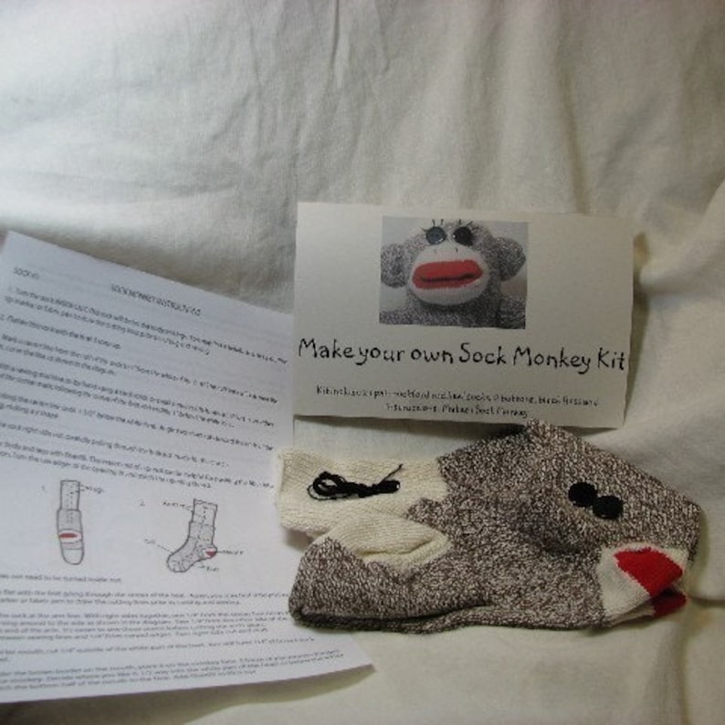 Puede incluir: Un kit de artesan&iacute;a de mono de calcet&iacute;n con un calcet&iacute;n marr&oacute;n y blanco, botones negros, hilo rojo e instrucciones sobre c&oacute;mo hacer tu propio mono de calcet&iacute;n. "Make your own Sock Monkey Kit" est&aacute; escrito en la tarjeta.