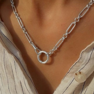 Peut inclure: Un collier à chaîne argentée composé de maillons ovales allongés et d'un fermoir circulaire proéminent au centre. Les maillons métalliques sont polis et réfléchissants, formant une boucle continue autour du cou, présentant un design classique.