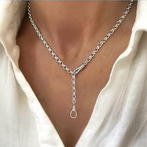 May include: A silver necklace with a Y-shaped design. The necklace has small, round links forming the chain. A clasp connects the chain, and a dangling pendant with a small loop at the end hangs from the centre.