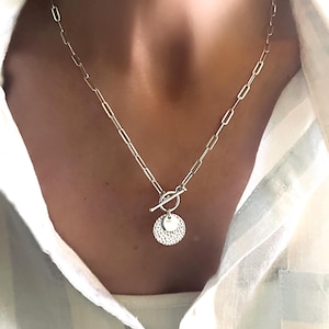 May include: Silver necklace with a toggle clasp and a pendant comprising a textured disc and a small heart charm. The chain has a link design. The necklace is worn against a white and light green striped shirt.