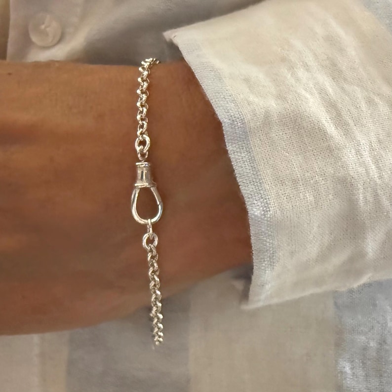 May include: A silver chain bracelet featuring a series of round, interlocking links and a central, elongated oval-shaped clasp mechanism. The bracelet is constructed from polished silver-toned metal, showcasing a classic link design with a secure closure.