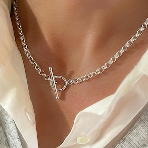 May include: A silver-toned chain necklace featuring a toggle clasp closure. The necklace is constructed from a series of interconnected round links, with a central T-bar and circular ring mechanism that serves as the functional fastening point.