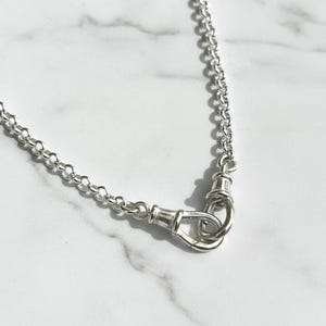 May include: A silver necklace with a delicate chain and a unique clasp. The clasp features a looped design, adding a touch of elegance. The necklace is displayed on a white marble surface, highlighting its shine.