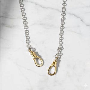 May include: Two silver-tone chain straps with gold-coloured lobster claw clasps. The chains are made of small, linked circles. The clasps are a shiny gold colour. The background is a white marble pattern.