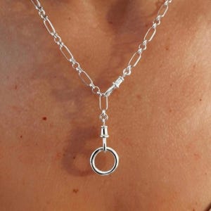 May include: A silver-toned necklace featuring a chain composed of elongated oval links. The necklace includes a central vertical connector piece that attaches to a circular ring pendant, creating a clean and metallic aesthetic design.