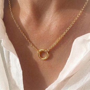 May include: A gold-coloured necklace featuring a delicate cable chain and a single, small, circular pendant ring at the centre. The chain links are uniform and round, connecting to the central ring, which rests against the skin of the neck.