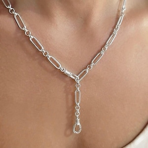May include: A silver-coloured necklace featuring elongated oval links connected by a central clasp. A short vertical drop chain extends from the clasp, ending in a small, U-shaped pendant component. The chain has a polished, reflective metallic finish.