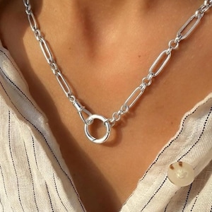 May include: A silver-toned necklace featuring elongated oval links connected to a central circular clasp. The chain is composed of polished metal links that create a uniform, repeating pattern around the neck, secured by a round spring-ring closure.