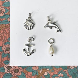 May include: Four silver charms: a seashell, a dolphin, an anchor, and a pearl. Each charm has a small loop for attaching to a bracelet or necklace. The charms are on a white surface with a floral patterned background.
