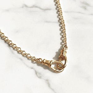 May include: A gold-toned metal chain necklace featuring a series of uniform circular links. The necklace is secured with a prominent, oval-shaped swivel clasp mechanism that connects the two ends of the chain together in a secure, interlocking loop.