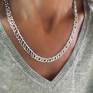 May include: A silver-colored chain necklace with a woven design. The necklace is worn around the neck and is made of a shiny metal. The background is a gray sweater.