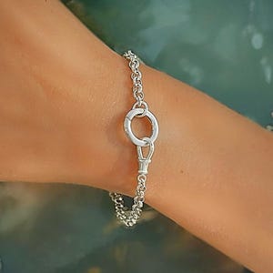 May include: A silver chain bracelet featuring a series of interlocking circular links and a prominent round spring ring clasp. The bracelet is constructed from polished metallic silver, showcasing a uniform link pattern that connects to a central circular closure mechanism.