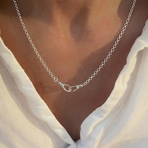 May include: A silver-coloured rolo chain necklace featuring a prominent front-facing clasp mechanism. The necklace is composed of uniform circular links that connect to a central hook-and-eye style closure, creating a continuous metallic loop design.