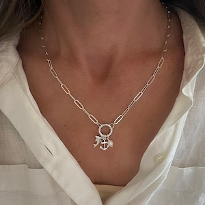 May include: A silver-colored necklace with a chain of elongated links. The necklace features a circular pendant with charms, including a dolphin, an anchor, and a small pearl. The necklace is worn against a white shirt.