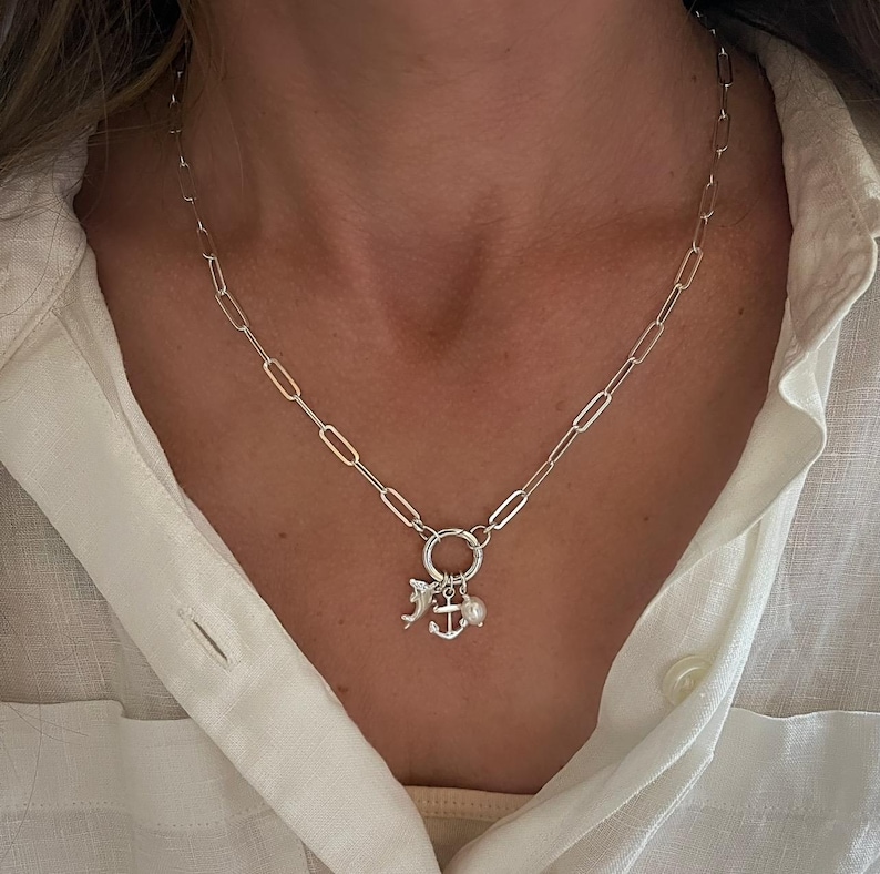 May include: A silver-colored necklace with a chain of elongated links. The necklace features a circular pendant with charms, including a dolphin, an anchor, and a small pearl. The necklace is worn against a white shirt.
