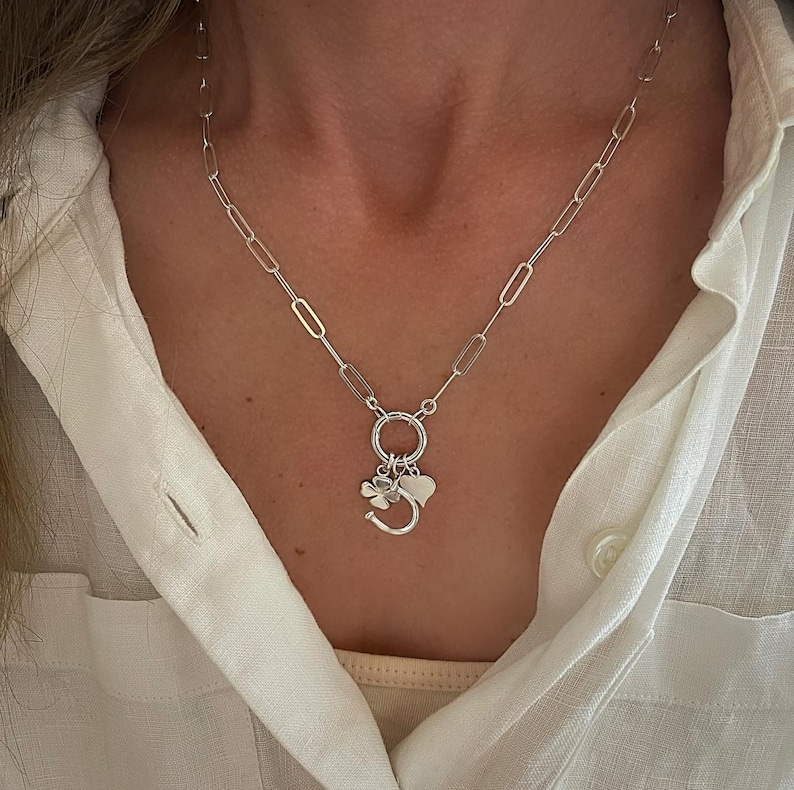 May include: A silver-colored necklace with a paperclip chain design. The necklace features a charm cluster with a horseshoe, a four-leaf clover, and a heart pendant. The necklace is worn against a white button-down shirt.