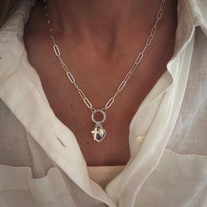 May include: A silver-toned necklace with a chain of elongated oval links. A small ring holds a heart-shaped charm, a cross, and a small pearl. The necklace is worn over a white button-down shirt.
