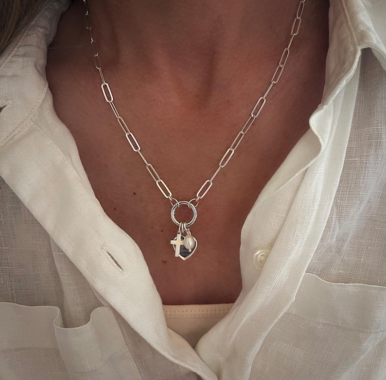 May include: A silver-toned necklace with a chain of elongated oval links. A small ring holds a heart-shaped charm, a cross, and a small pearl. The necklace is worn over a white button-down shirt.