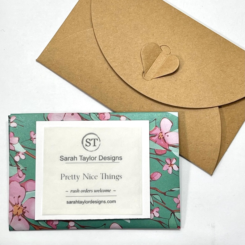 May include: A rectangular card featuring a teal background with pink floral patterns, overlaid with a white square label displaying the text Sarah Taylor Designs, Pretty Nice Things, and sarahtaylordesigns.com, accompanied by a brown paper envelope.