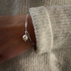 May include: A silver chain bracelet featuring elongated rectangular links and a single round white pearl charm. The bracelet is secured with a metal jump ring, showcasing a polished metallic finish and a smooth, lustrous surface on the pearl.