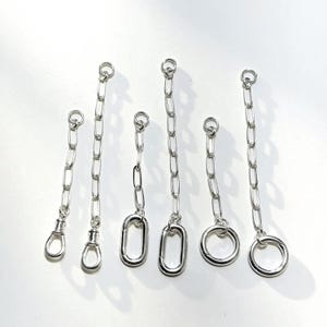 May include: A selection of silver chain extenders with different end designs. The extenders have small chains with a loop at one end and a clasp, oval, or ring at the other. They are presented on a white background.