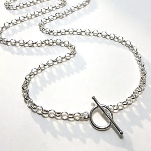 May include: A silver-colored necklace with a toggle clasp. The chain is made of small, round links. The clasp features a circular ring and a straight bar. The necklace is displayed on a white surface.
