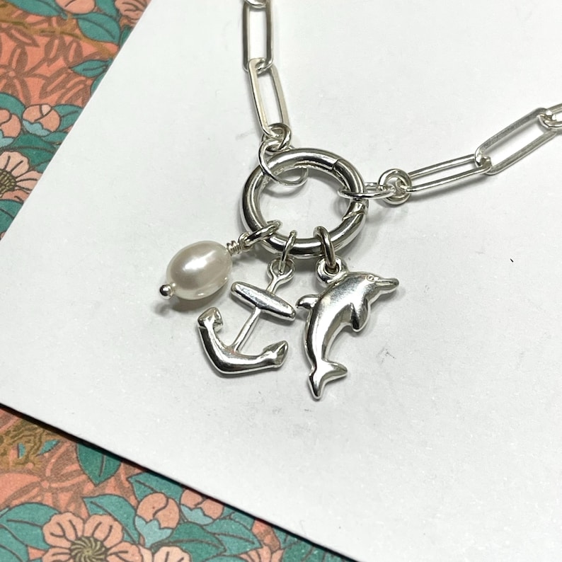 May include: Silver-toned necklace featuring a large circular clasp and a chain with elongated links. The necklace has three charms: a pearl, an anchor, and a dolphin. The necklace is displayed on a white surface.