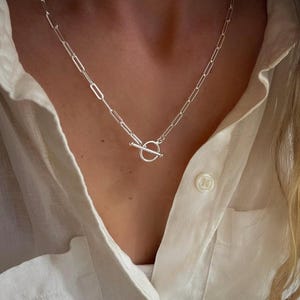 May include: A silver-toned chain necklace featuring elongated rectangular links and a circular toggle clasp closure. The necklace is worn around the neck, resting against a white collared shirt with a visible button detail near the neckline.