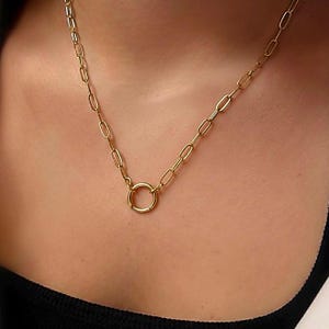 May include: A gold necklace featuring a circular pendant. The chain is made of oval-shaped links. The pendant is a plain, smooth circle. The necklace is worn on a person's neck, against a dark top. The chain links are oval-shaped.