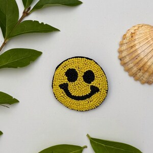 Peut inclure: Un écusson jaune en forme de smiley avec des yeux noirs et une bouche incurvée. Un coquillage et des feuilles vertes sont également présents dans l'image.