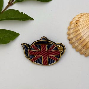 Pode incluir: Broche decorativo em forma de bule com design da Union Jack em vermelho, azul e dourado. Colocado sobre um fundo branco com uma concha e folhas verdes. Um acessório de moda ou lembrança.