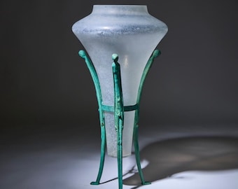 May include: A frosted glass vase with a flared top sits on a green metal stand with four legs shaped like human figures.