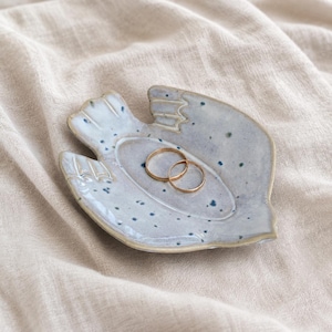 May include: A light blue ceramic trinket dish shaped like a bird with dark blue speckles throughout the surface. Two thin gold-toned metal rings are resting in the center of the dish, which features defined wing and tail details.