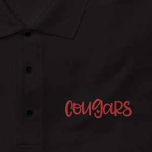 Puede incluir: Polo negro con un logotipo bordado en rojo "Cougars" en el lado derecho. La camisa tiene una etiqueta azul que dice "Port Authority".