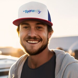 May include: A man wearing a white and blue baseball cap with the word "howdy" on it. He is smiling and wearing a gray hoodie over a black t-shirt.