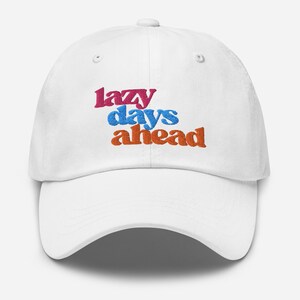 Puede incluir: Gorra de béisbol blanca con la frase bordada "lazy days ahead" en rosa, azul y naranja. La gorra tiene una visera curva y una correa ajustable. Un accesorio casual para el uso diario.